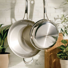 Two EverLast Tri-Ply Clad Stainless Steel Frying Pan Set pans hang on a tiled kitchen wall, with green potted plants and a wooden cutting board nearby, highlighting stylish cookware in a cozy setting.
