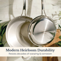 Two EverLast Tri-Ply Clad Stainless Steel Frying Pans hang on a tiled kitchen wall with green plants and a wooden cutting board. Text reads: Frying Pan Set—Modern Heirloom Durability that resists decades of warping & corrosion.