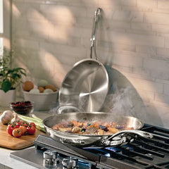 An EverLast Tri-Ply Clad Stainless Steel Frying Pan Set rests on a gas stove with food and rising steam, while another pan from the set shines in the background surrounded by fresh vegetables, eggs, and kitchen essentials.