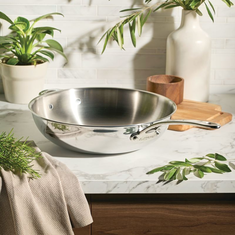 The EverLast Tri-Ply Clad Stainless Steel 12.25-Inch Stir Fry with Helper Handle sits on a marble countertop, surrounded by plants and kitchen accessories—perfect cookware for the modern home chef in a sunlit, inviting space.