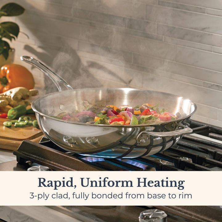 The EverLast Tri-Ply Clad Stainless Steel 12.25-Inch Stir Fry with Helper Handle cooks veggies on a gas stove, steaming. Rapid, uniform heating—3-ply clad from base to rim—makes it ideal for any home chef.