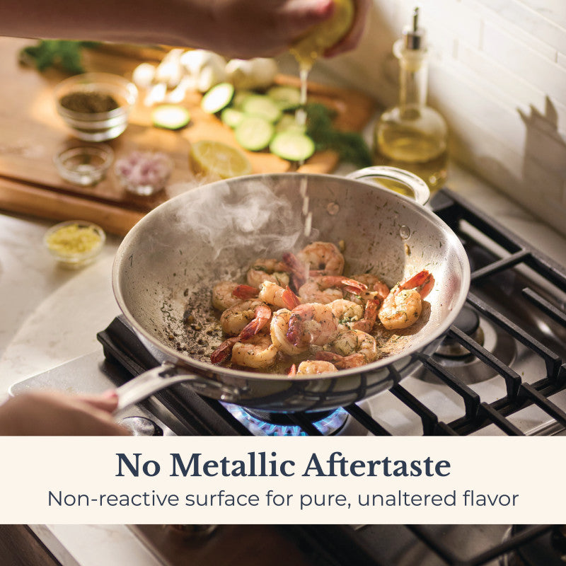 A hand squeezes lemon over shrimp in an EverLast Tri-Ply Clad Stainless Steel 12.25-Inch Stir Fry with Helper Handle. Cucumber and garlic are on the counter. Text: No Metallic Aftertaste—non-reactive surface preserves pure, unaltered flavor.