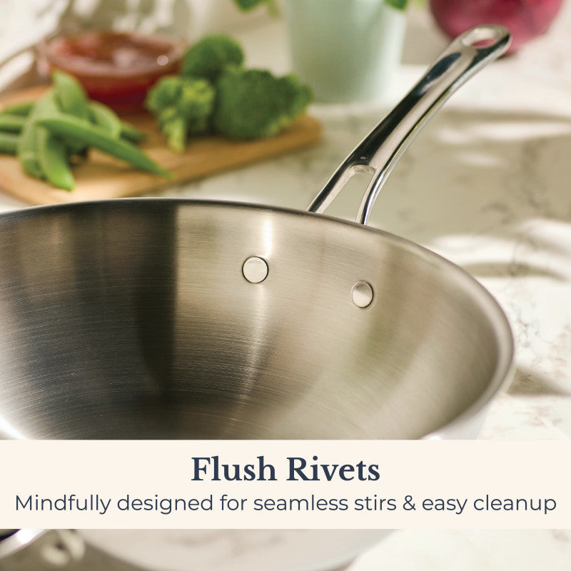 An EverLast Tri-Ply Clad Stainless Steel 12.25-Inch Stir Fry with Helper Handle sits on a marble countertop; blurred fresh vegetables and a cutting board are in the background. Text: Flush Rivets—Designed for seamless stirs & easy cleanup.