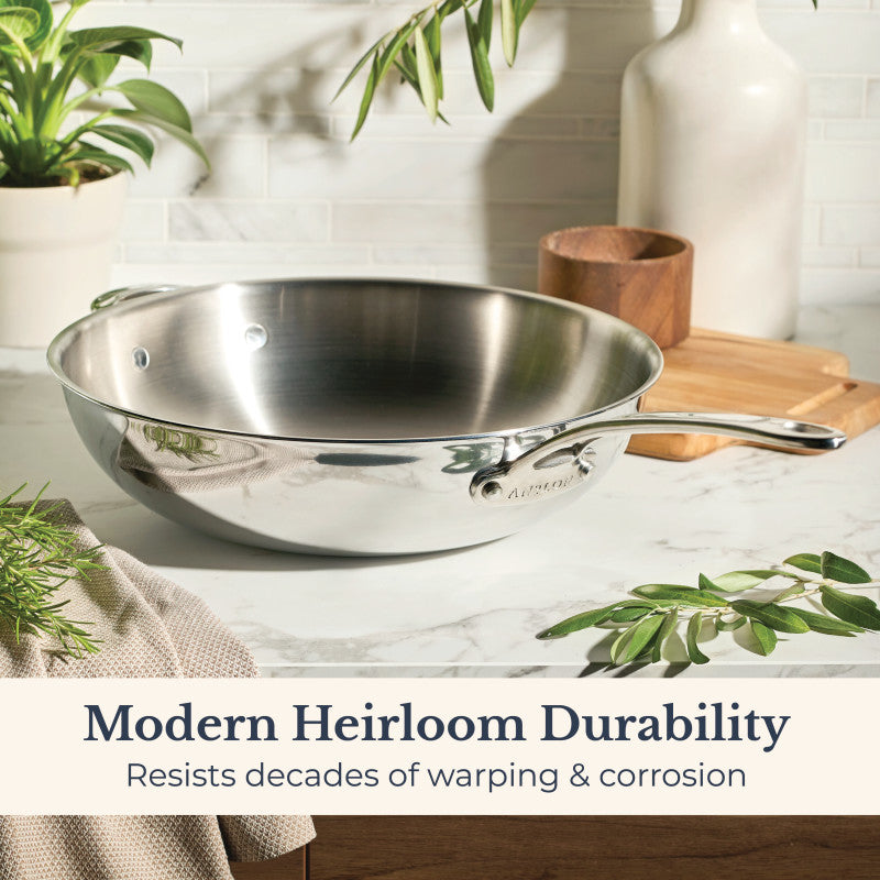 An EverLast Tri-Ply Clad Stainless Steel 12.25-Inch Stir Fry with Helper Handle rests on a marble counter beside plants, a napkin, and wooden container. Text: “Modern Heirloom Durability—resists warping & corrosion for decades.”.
