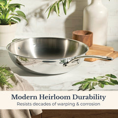 An EverLast Tri-Ply Clad Stainless Steel 12.25-Inch Stir Fry with Helper Handle rests on a marble counter beside plants, a napkin, and wooden container. Text: “Modern Heirloom Durability—resists warping & corrosion for decades.”.