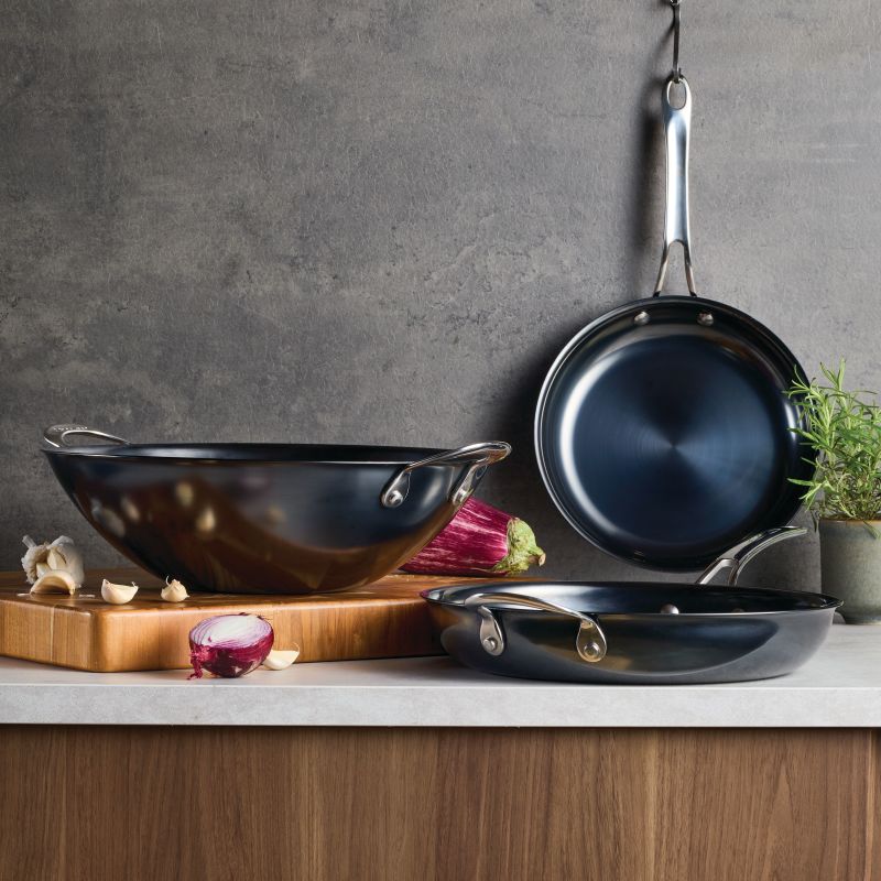 Three N₂ Carbon Steel 10-Inch Frying Pans are shown on a kitchen counter: one hangs on the wall, one sits flat, and another rests on a wooden cutting board with garlic, onion, eggplant, and rosemary nearby.
