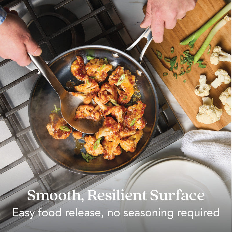 A person cooks cauliflower in an N₂ Carbon Steel 10-Inch Frying Pan using a spatula and spoon. Chopped green onions and raw cauliflower are nearby. Text reads: Smooth, resilient surface. Rust resistant with easy food release—no seasoning needed.