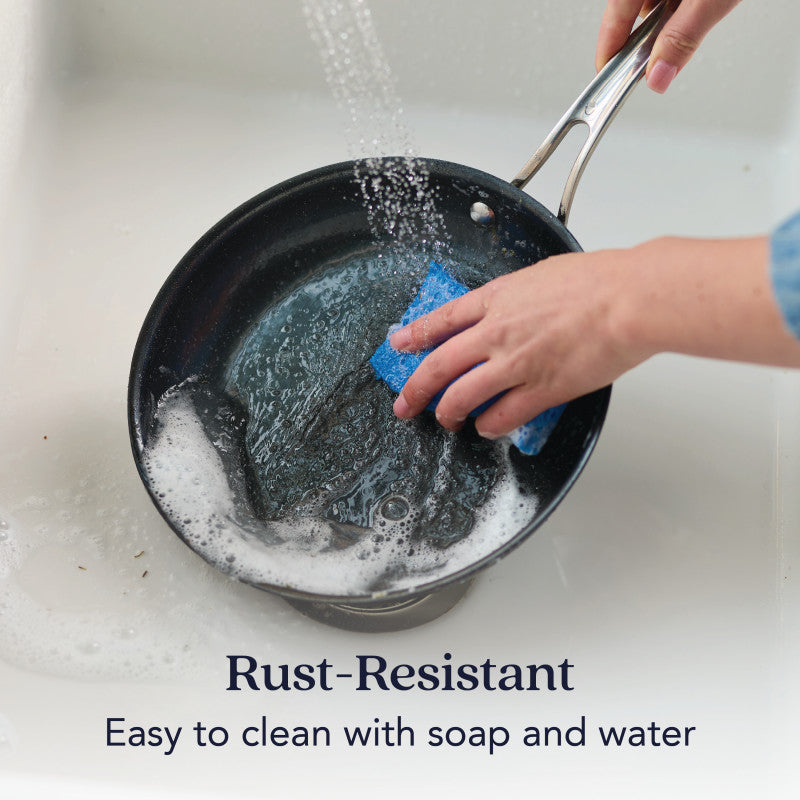 Someone washes the 10-Inch N₂ Carbon Steel Frying Pan with a blue sponge under running water. Text reads: Rust-resistant. Easy to clean with soap and water while preserving its nonstick patina. Brand: N₂ Carbon Steel.