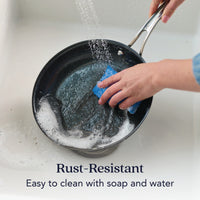 Someone washes the 10-Inch N₂ Carbon Steel Frying Pan with a blue sponge under running water. Text reads: Rust-resistant. Easy to clean with soap and water while preserving its nonstick patina. Brand: N₂ Carbon Steel.