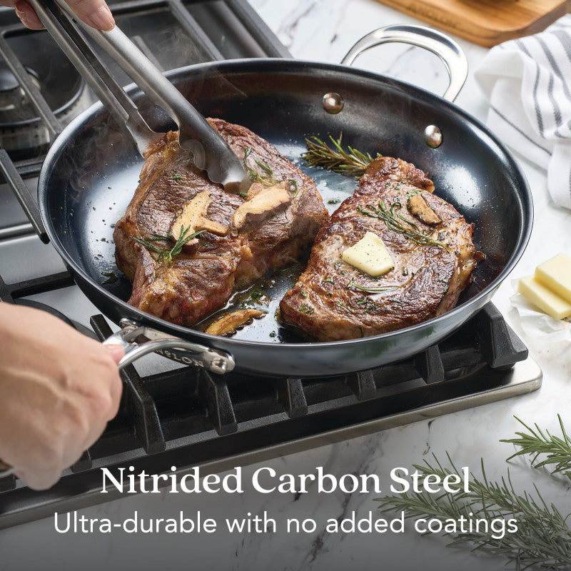 Two steaks with butter and rosemary sizzle in a black skillet on a gas stove as a hand turns one with tongs. Text reads: N₂ Carbon Steel 12-Inch Frying Pan—ultra-durable, naturally nonstick, no added coatings.