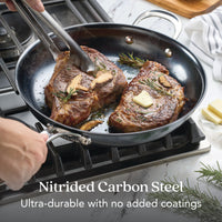 Two steaks with butter and rosemary sizzle in a black skillet on a gas stove as a hand turns one with tongs. Text reads: N₂ Carbon Steel 12-Inch Frying Pan—ultra-durable, naturally nonstick, no added coatings.