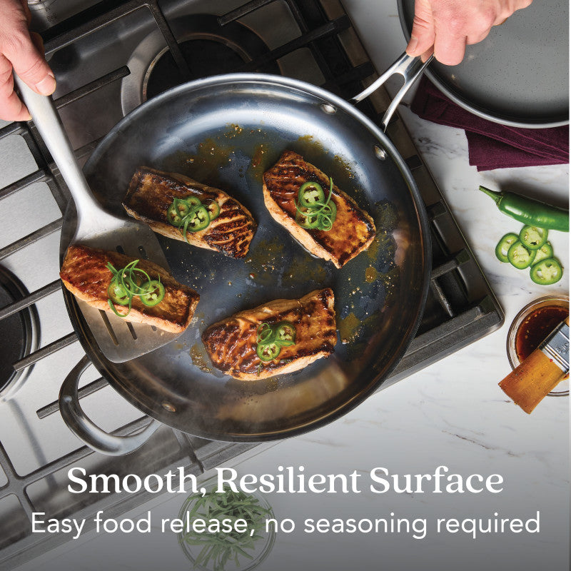 A person lifts a seared fish fillet from the 12-Inch N₂ Carbon Steel Frying Pan on a stove, while three fillets with sliced green chilies remain. Naturally evolving nonstick surface: easy food release, no seasoning required.