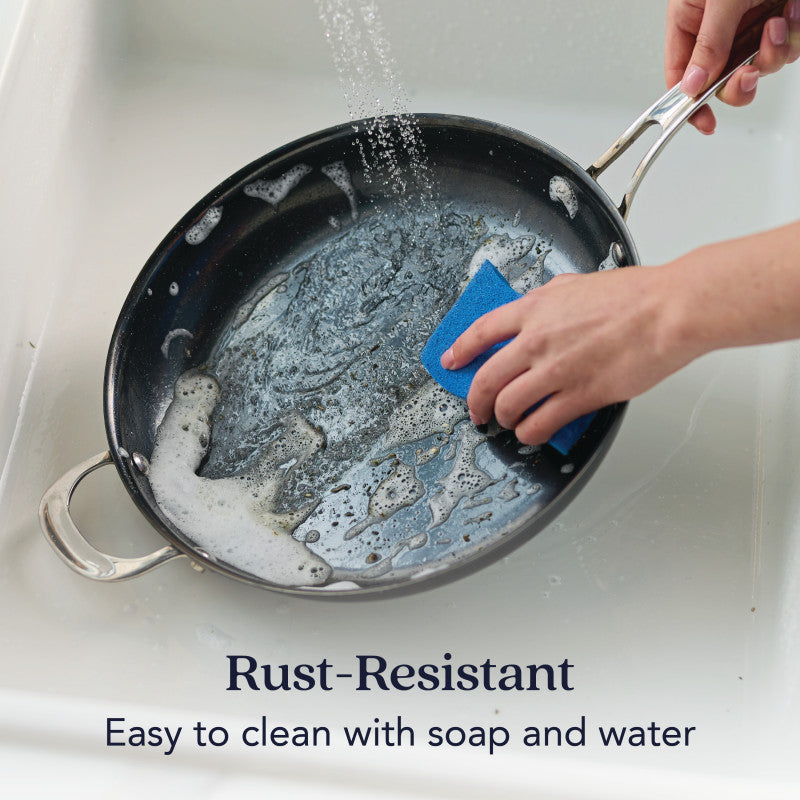 A hand scrubs the N₂ Carbon Steel 12-Inch Frying Pan with a blue sponge under running water. Text: Rust-resistant due to advanced nitriding technology. Easy to clean with soap and water. Brand: N₂ Carbon Steel.