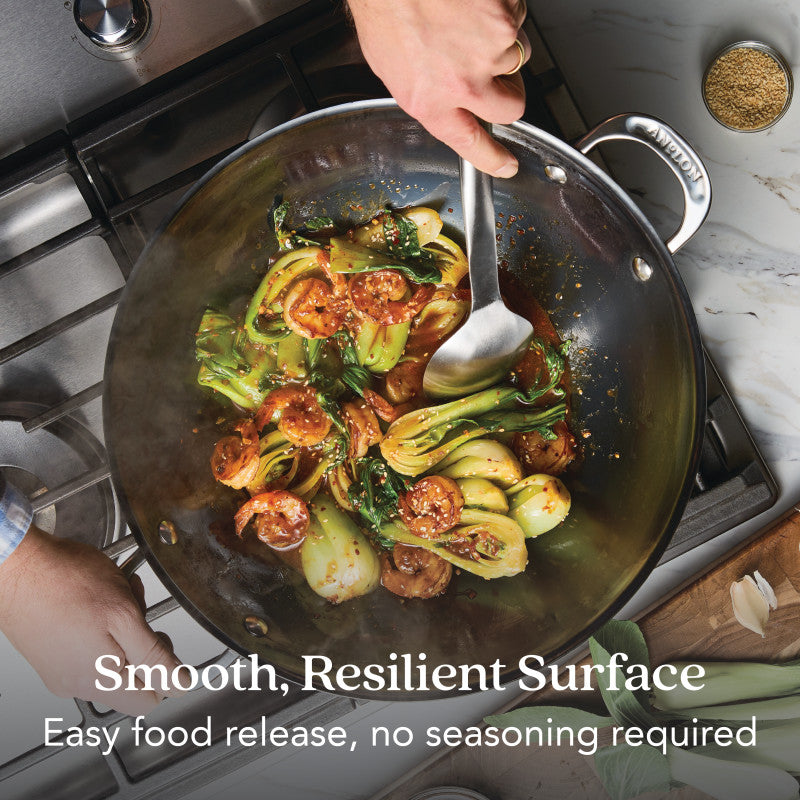 A person stirs bok choy and sausage in an N2 Carbon Steel Wok by N2 Carbon Steel on a stovetop. Text reads: Smooth, resilient surface—nitriding technology for easy food release, no seasoning needed. Utensils and a bowl of pepper sit nearby.