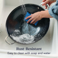A person scrubs a black N2 Carbon Steel Wok with a blue sponge under running water. Soap suds are visible. Text: Rust-resistant N2 Carbon Steel cookware—easy to clean with soap and water.