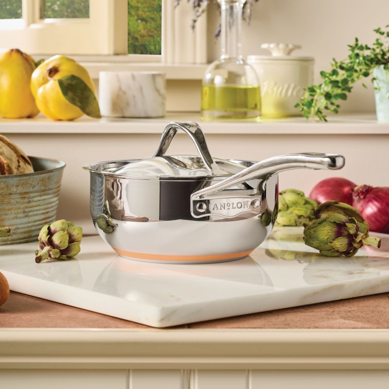 A 2.5-Quart Saucier with Lid from Nouvelle Stainless sits on a white marble cutting board in a kitchen, surrounded by artichokes, onions, and other fresh produce.
