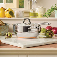 A 2.5-Quart Saucier with Lid from Nouvelle Stainless sits on a white marble cutting board in a kitchen, surrounded by artichokes, onions, and other fresh produce.