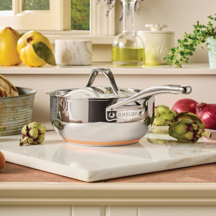 A 2.5-Quart Saucier with Lid from Nouvelle Stainless sits on a white marble cutting board in a kitchen, surrounded by artichokes, onions, and other fresh produce.