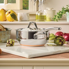 A 2.5-Quart Saucier with Lid from Nouvelle Stainless sits on a white marble cutting board in a kitchen, surrounded by artichokes, onions, and other fresh produce.