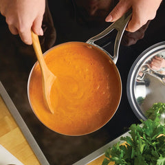 A person stirs creamy orange soup on a stovetop with a wooden spoon in a Nouvelle Stainless 2.5-Quart Saucier with Lid; a metal lid rests nearby, and fresh herbs are visible in the corner.