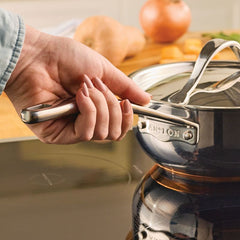 A hand holds the handle of a Nouvelle Stainless 2.5-Quart Saucier with Lid on a stovetop; blurred vegetables in the background highlight its optimum heat control.
