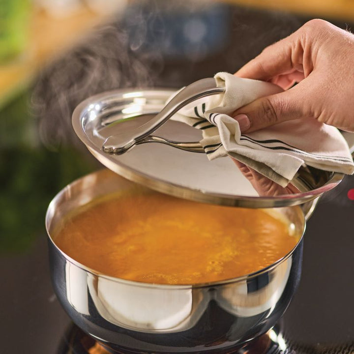 A hand uses a cloth to lift the lid from a steaming pot of soup simmering in a Nouvelle Stainless 2.5-Quart Saucier with Lid on the stovetop.