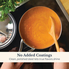 A hand stirs orange soup in a Nouvelle Stainless 2.5-Quart Saucier with Lid. Clean, polished steel ensures optimum heat control and pure flavor, with no added coatings. Fresh herbs and the lid are nearby.