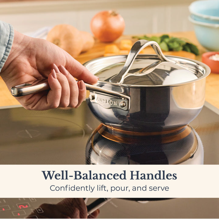 A hand lifts the Nouvelle Stainless 2.5-Quart Saucier with Lid by its handle on an induction stove. Text: Well-Balanced Handles—lift, pour, and serve with confidence.