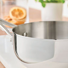 A Nouvelle Stainless 2.5-Quart Saucier with Lid gleams on a white countertop, its handle visible. In the blurred background sit a potted plant and a glass of water with dried orange slices, hinting at precise heat control for perfect results.