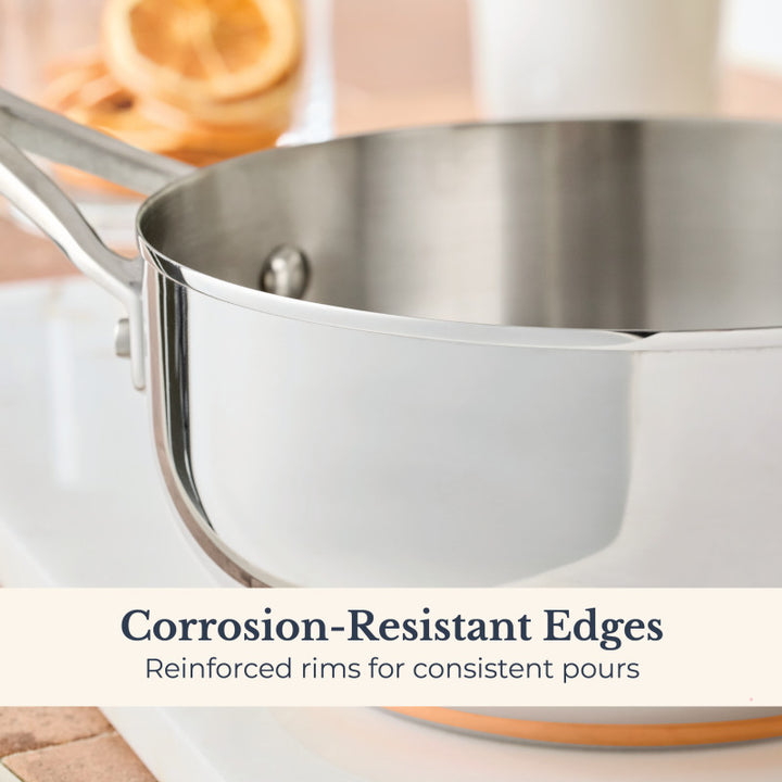 A close-up of a Nouvelle Stainless 2.5-Quart Saucier with Lid showcases corrosion-resistant reinforced rims for consistent pours. Blurred dried orange slices appear in the background.