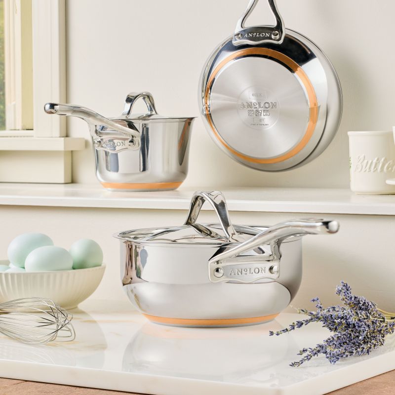 A Nouvelle Stainless 2.5-Quart Saucier with Lid rests on a white countertop near blue eggs, a whisk, and lavender; another saucier and lid hang on the wall above.