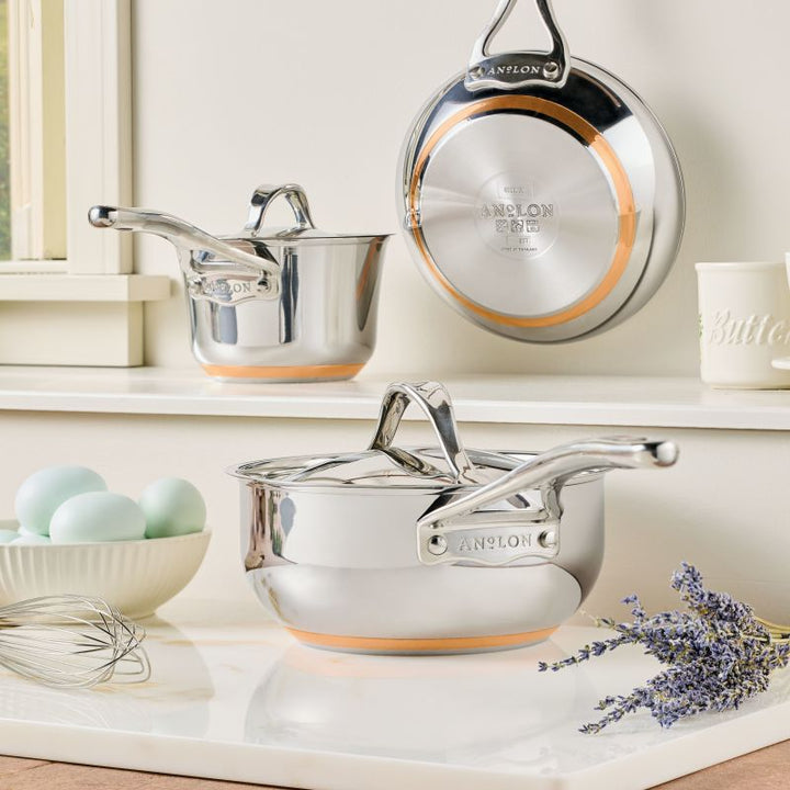 A Nouvelle Stainless 2.5-Quart Saucier with Lid rests on a white countertop near blue eggs, a whisk, and lavender; another saucier and lid hang on the wall above.