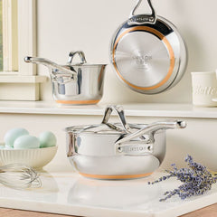 A Nouvelle Stainless 2.5-Quart Saucier with Lid rests on a white countertop near blue eggs, a whisk, and lavender; another saucier and lid hang on the wall above.