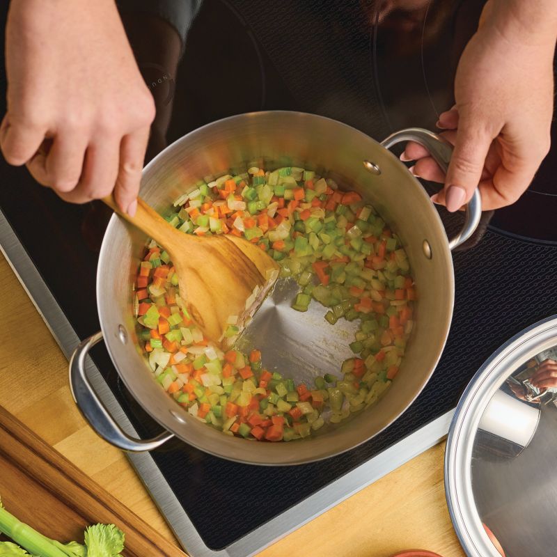 A person stirs chopped celery, carrots, and onions in a pot on the stove with a wooden spatula. Nearby is a Nouvelle Stainless 6.5-Quart Stockpot with Lid and assorted cookware, along with fresh vegetables.