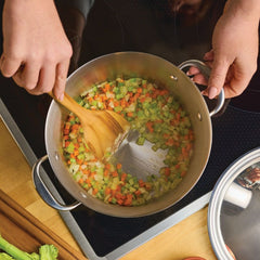A person stirs chopped celery, carrots, and onions in a pot on the stove with a wooden spatula. Nearby is a Nouvelle Stainless 6.5-Quart Stockpot with Lid and assorted cookware, along with fresh vegetables.