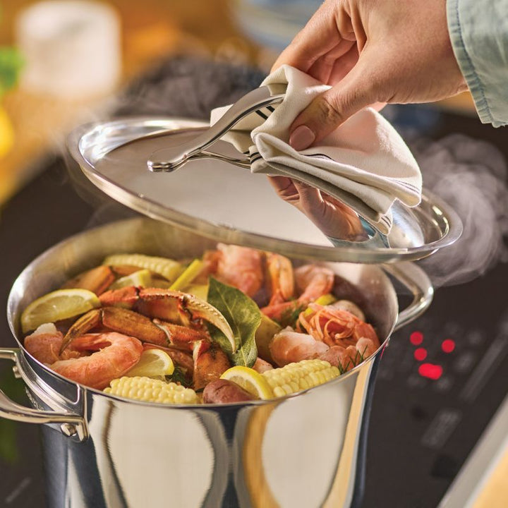 A hand lifts the lid off a Nouvelle Stainless 6.5-Quart Stockpot on a stove, revealing a seafood boil with shrimp, crab legs, corn on the cob, lemon slices, and herbs.