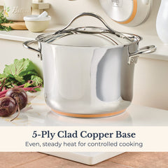 A Nouvelle Stainless 6.5-Quart Stockpot with Lid sits on a white surface next to purple onions and green leafy veggies. Text reads: “5-Ply Clad Copper Base. Even, steady heat for controlled cooking.”.