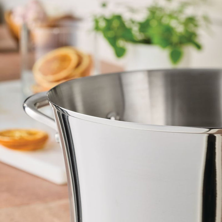 A close-up of the Nouvelle Stainless 6.5-Quart Stockpot with Lid, featuring sturdy handles, sits on a kitchen counter. Blurred in the background are glass jars, dried orange slices, and a green potted plant.