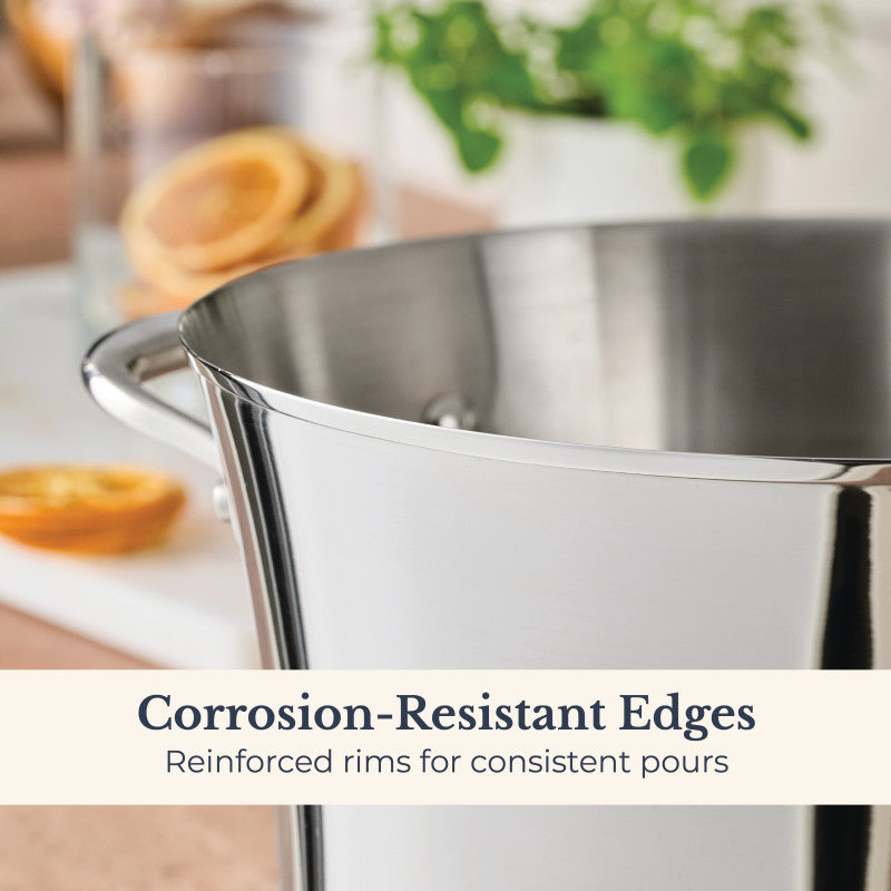 A close-up of the Nouvelle Stainless 6.5-Quart Stockpot with Lid, featuring reinforced, corrosion-resistant rims for consistent pours. In the blurred background: a cutting board with dried orange slices and green leaves. Premium cookware quality.