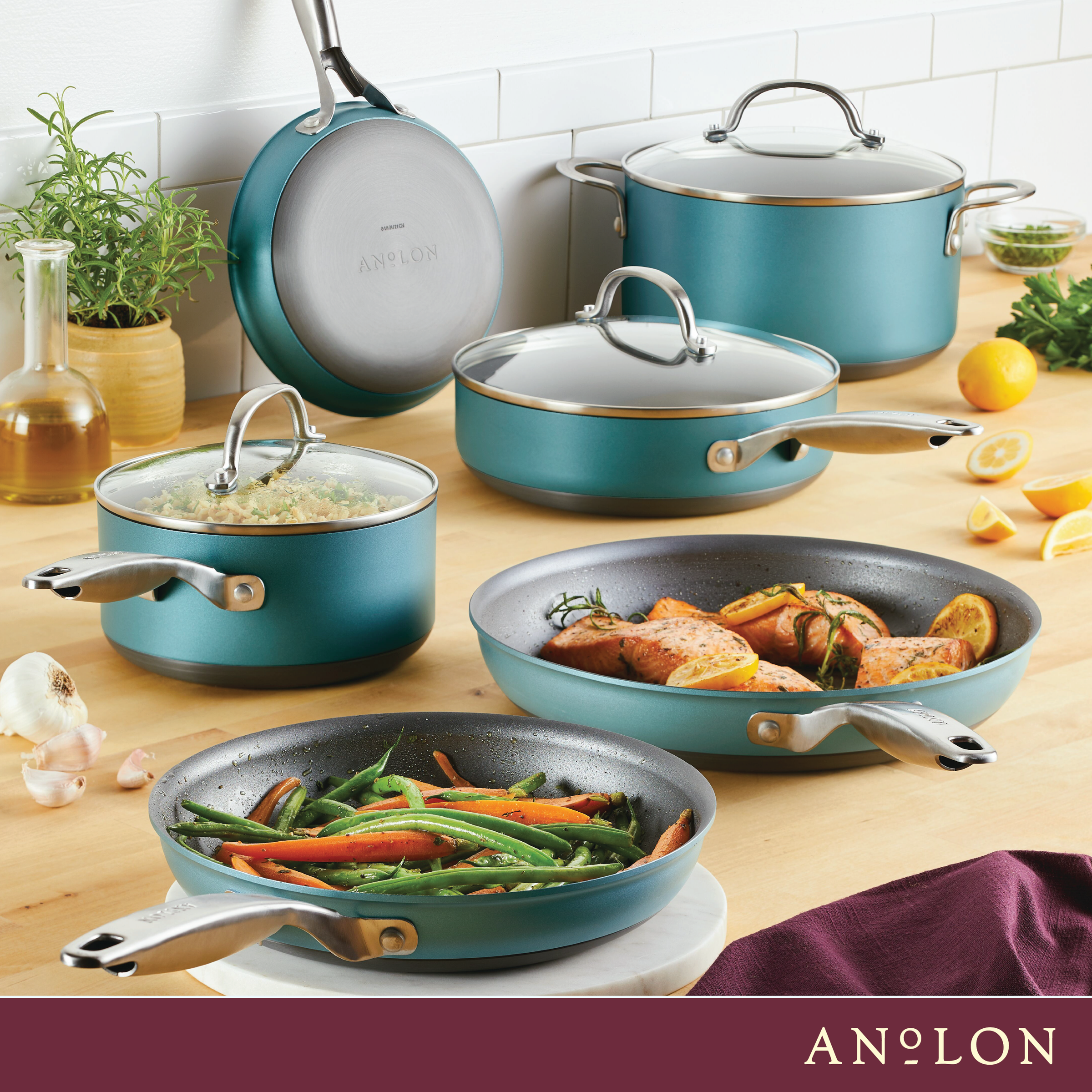 A collection of teal cookware, including pots and pans with glass lids, displayed on a kitchen counter. Vegetables and chicken are cooking in some pans. Lemons, garlic, a towel, and a plant are also on the counter.