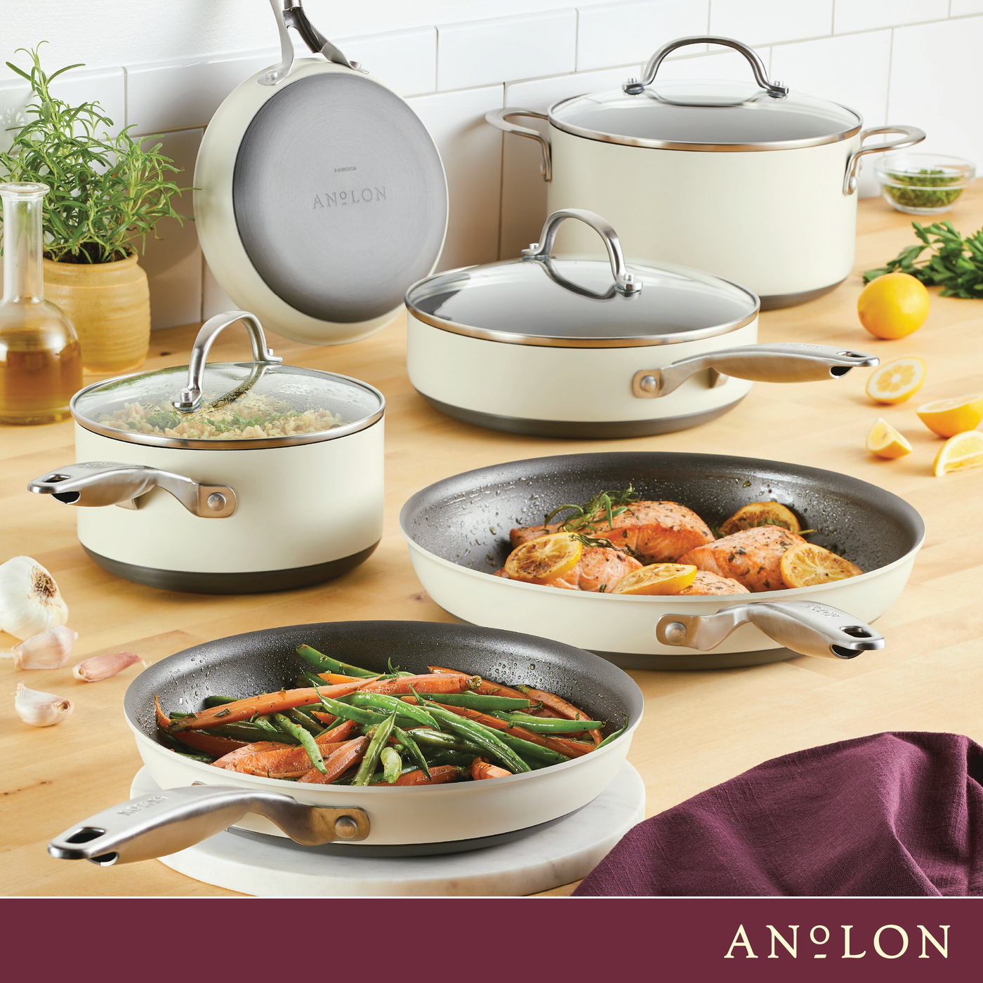 A kitchen scene with various Anolon cookware, including saucepans and frying pans. The pans contain cooked vegetables and salmon with lemon. Fresh herbs, garlic, and lemons are visible on the wooden countertop.