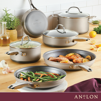 A kitchen scene with several Anolon cookware pieces on a wooden countertop. Pots and pans contain vegetables and a dish with chicken and roasted potatoes. Ingredients like garlic, oil, and lemons are nearby.
