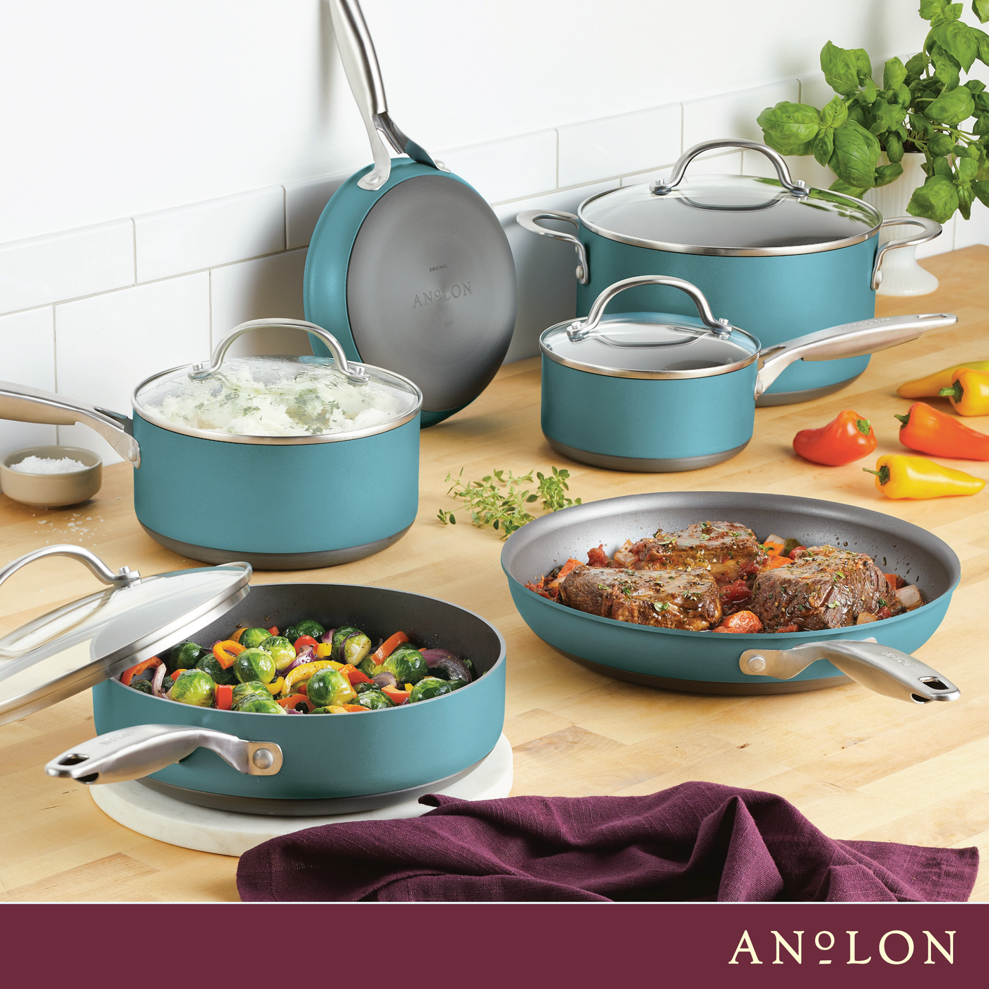 A kitchen scene with various teal pots and pans from Anolon, containing foods like mashed potatoes, beef, and vegetables. The cookware is arranged on a wooden counter, with colorful bell peppers and herbs in the background.
