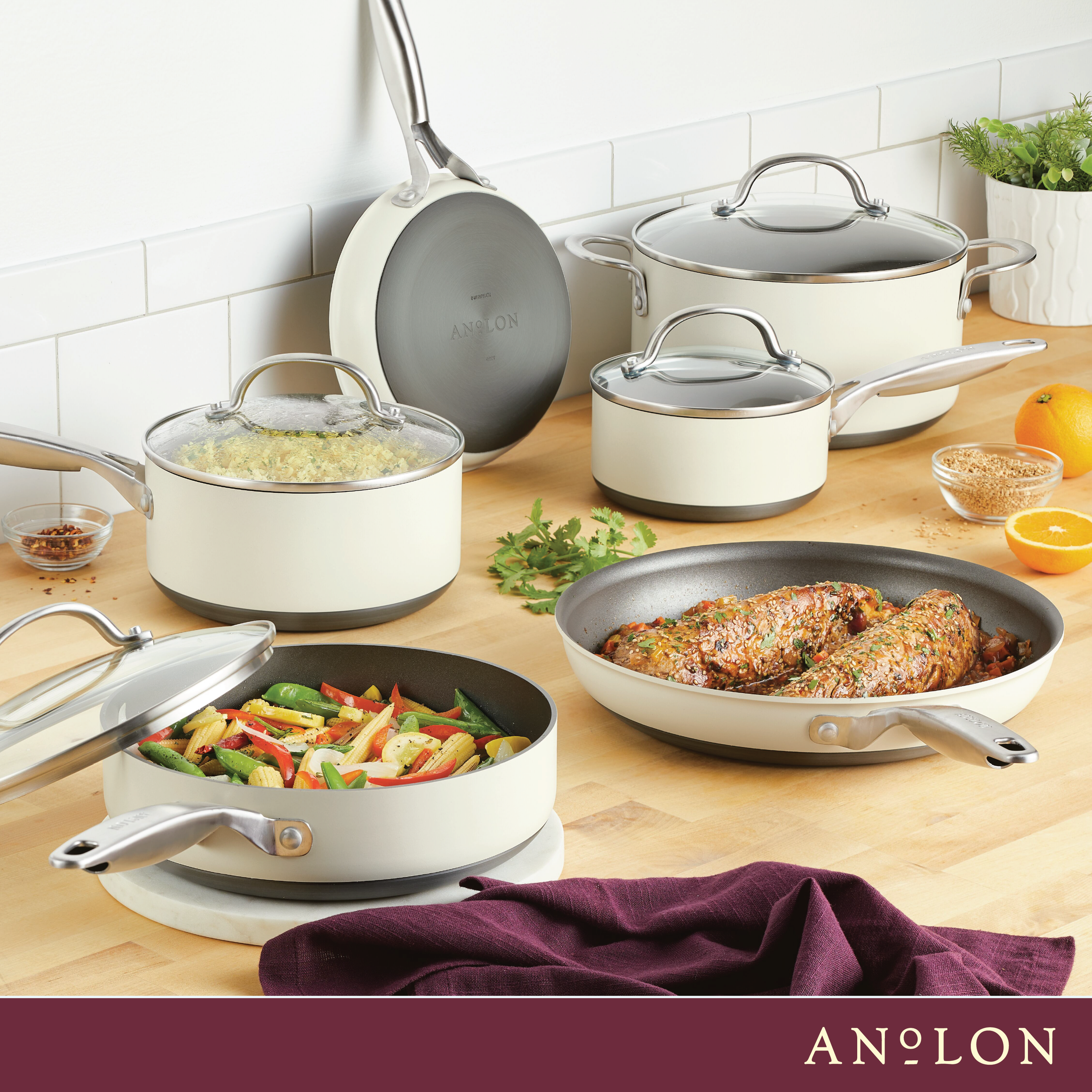 A kitchen scene featuring various Anolon pots and pans. Dishes include cooked vegetables in a frying pan and stuffed meat in a skillet. Other pots contain rice and are placed on a wooden countertop decorated with fresh herbs and citrus fruits.