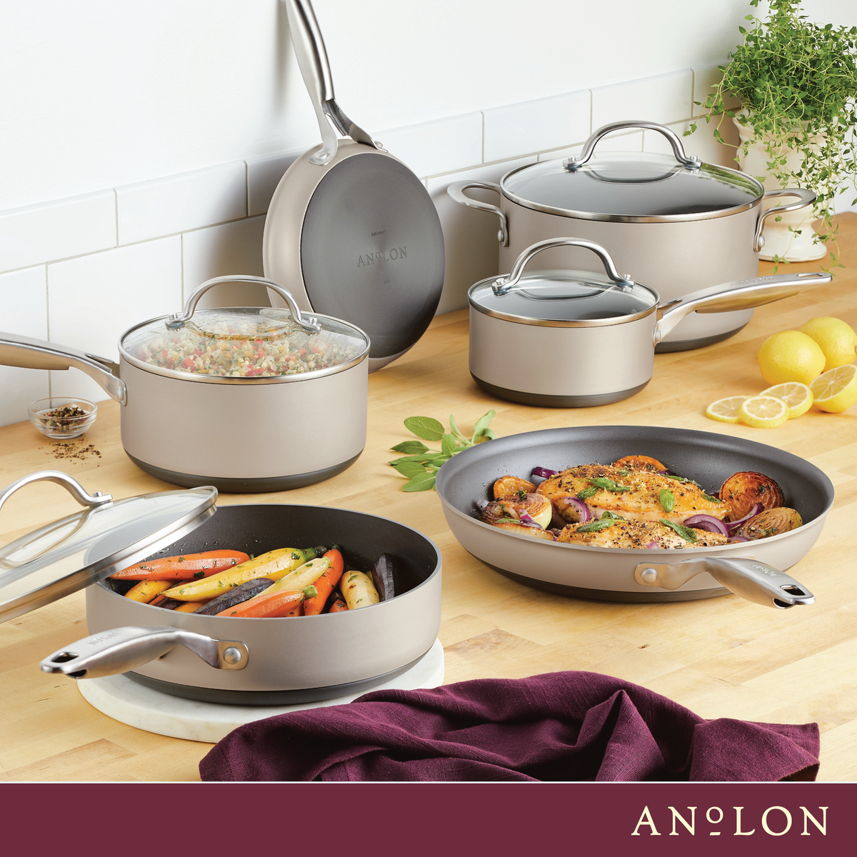 A kitchen scene displaying a set of various sized, non-stick pots and pans, some with glass lids. The cookware is silver-toned, and some contain cooked vegetables and chicken. Lemons and herbs are in the background, with a purple cloth in the foreground.