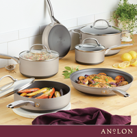 A kitchen scene displaying a set of various sized, non-stick pots and pans, some with glass lids. The cookware is silver-toned, and some contain cooked vegetables and chicken. Lemons and herbs are in the background, with a purple cloth in the foreground.