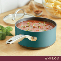 A teal saucepan filled with tomato sauce garnished with herbs sits on a wooden counter. In the background, theres a pot lid, fresh basil, and a bowl of uncooked pasta. The word Anolon is printed at the bottom of the image.