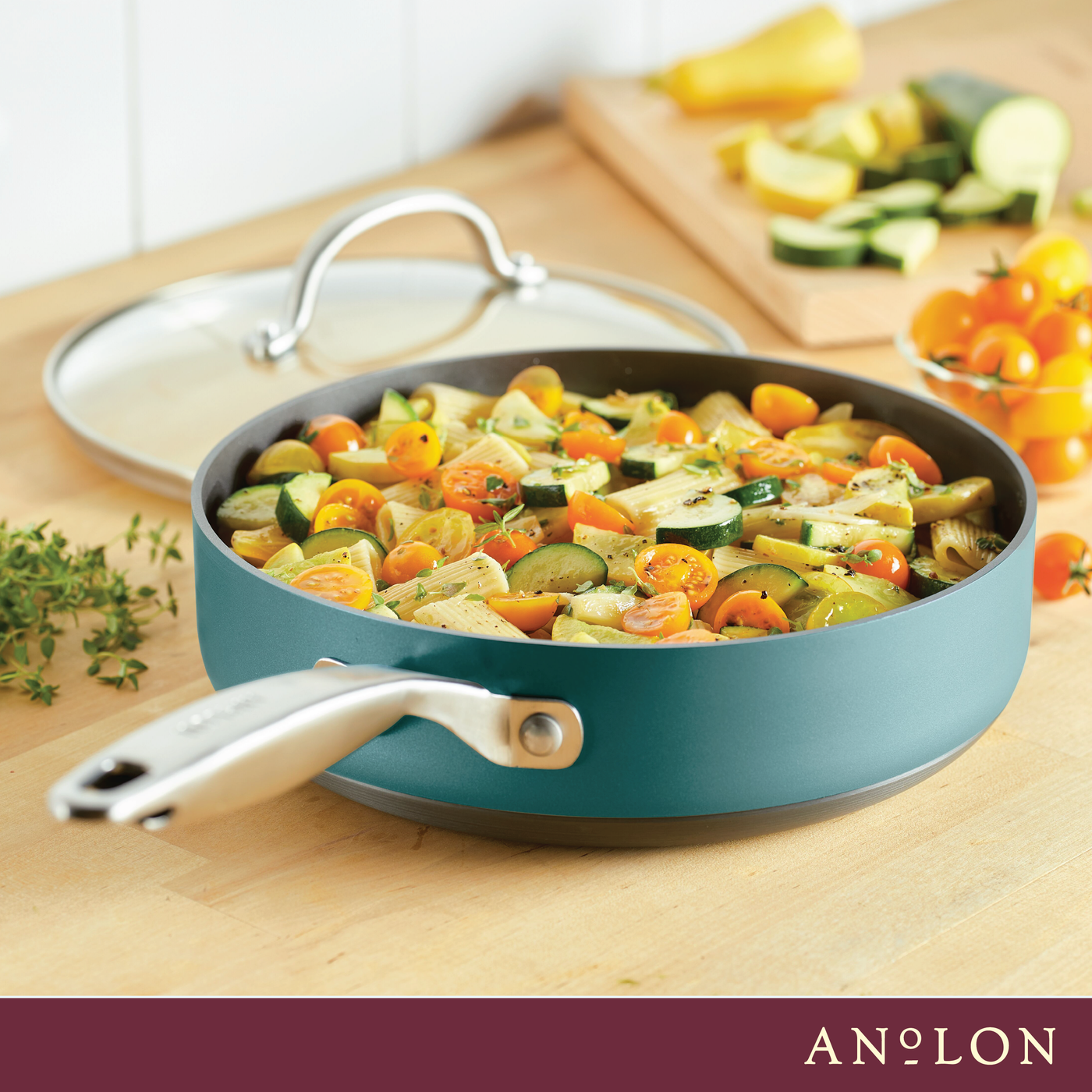 A turquoise frying pan filled with a colorful sauté of cherry tomatoes, zucchini, and squash sits on a wooden countertop. Fresh herbs are sprinkled on top, with a sprig of thyme and a lid nearby. A simple, elegant kitchen setting.