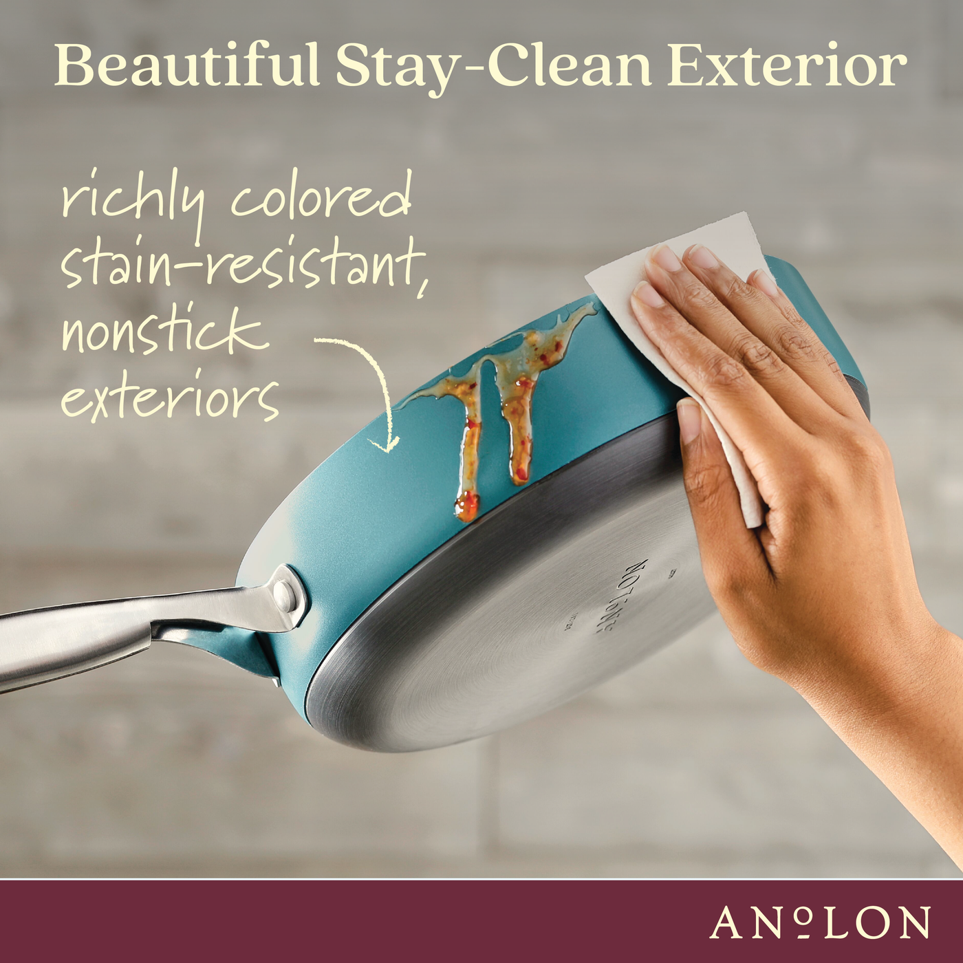 A hand wiping sauce off the exterior of a nonstick pan with a cloth. The pan is blue with a silver base, featuring the text Beautiful Stay-Clean Exterior and richly colored stain-resistant, nonstick exteriors. The brand name Anolon is visible.