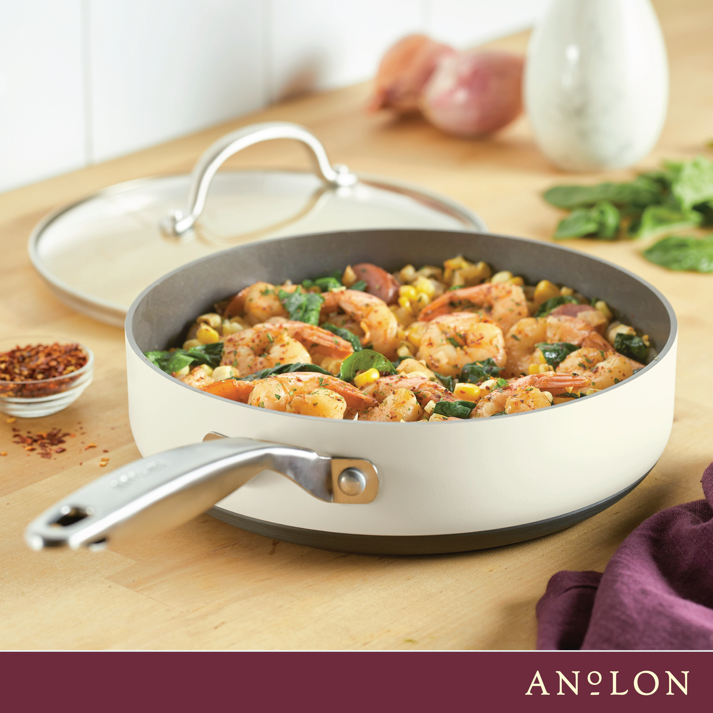 A skillet filled with cooked shrimp, corn, and vegetables sits on a kitchen counter. A glass lid rests nearby. A container of red pepper flakes and some fresh herbs are in the background.